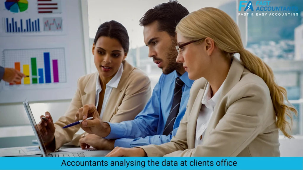 Three business professionals—two women and one man—sitting at a desk in a modern office, analyzing data on a laptop. A chart board with colorful graphs is visible in the background, indicating a client meeting or financial discussion. The group appears focused and collaborative.